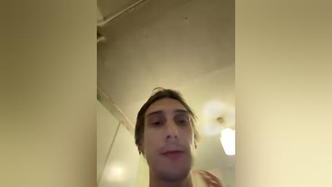 Video of a young man with light skin and brown hair, looking slightly down, standing in a dimly lit room with beige walls and a ceiling light.