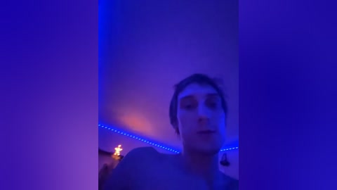 A video of a young man with short dark hair, captured in a dimly lit room with purple lighting. He has a neutral expression and is wearing a dark shirt. The background includes a wall with blue LED lights and a fireplace with glowing coals.