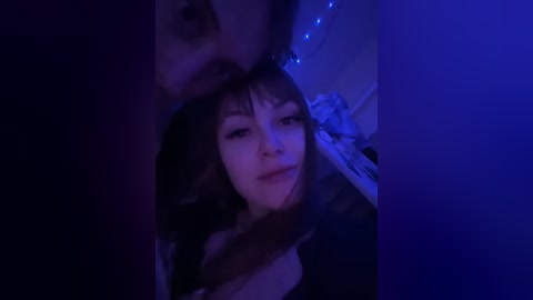 A dimly-lit video featuring a young woman with shoulder-length brown hair, smiling warmly, partially obscured by a man's face. Background includes a blue-toned room with soft lighting and a bed with rumpled white sheets.