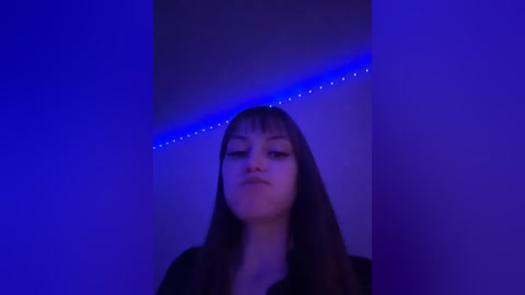 Video of a young woman with long dark hair and light skin, wearing a black top, standing under a blue LED strip light, creating a surreal, moody atmosphere.