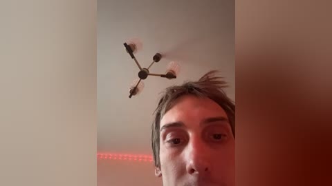 Video of a young man with light brown hair, partially visible, looking up at a drone with four propellers spinning, in a dimly lit room with red LED strip lighting.