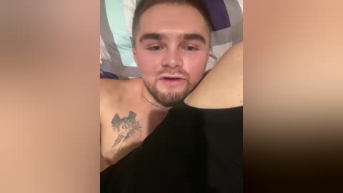 A video of a shirtless man with a beard, a tattoo of a wolf on his shoulder, and a man's legs in the foreground. He is lying on a bed with a striped blanket.