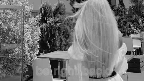 Black-and-white video of a woman with long, straight hair, wearing an off-shoulder dress, standing in a modern outdoor dining area with glass walls, surrounded by greenery and trees.