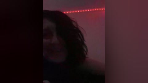 A dimly lit video of a person with messy black hair, partially obscured by a dark object, with a red LED strip light in the background.