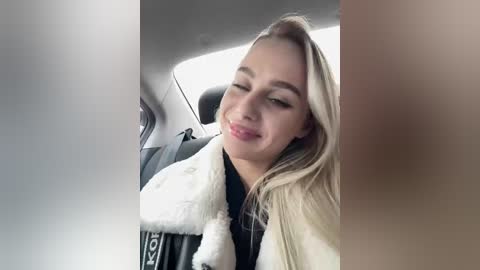 Video of a smiling young blonde woman in a car, wearing a white fur jacket and black top, with blurred window and seat visible in the background.