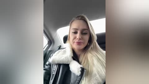 Video of a smiling, fair-skinned woman with long blonde hair, wearing a white fur-trimmed black leather jacket, seated in a car.