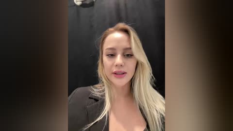 Video of a fair-skinned, blonde woman with long hair, wearing a black jacket, in a dimly lit, black-walled room. She has a neutral expression and appears to be mid-sentence.