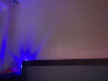 Video of a dimly lit room with a large bed and soft purple lighting. The bed has a grey headboard and a dark wooden frame, with a white curtain partially visible on the left. The background is dark, creating a moody atmosphere.