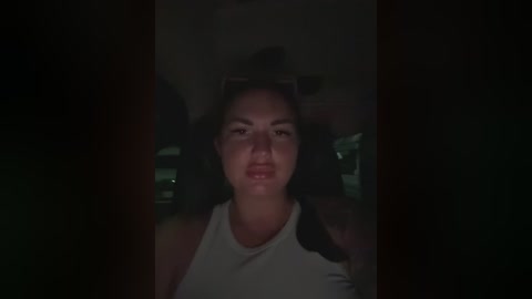 A dimly lit video of a young woman with light skin and dark hair, wearing a white tank top, in a dimly lit room, likely taken with a low-quality camera.
