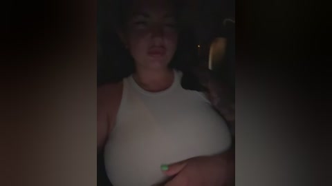 A dimly lit video shows a curvy woman with light skin, wearing a tight white tank top, partially visible breasts, and a green-painted fingernail, taken indoors.