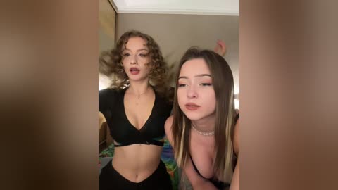 A video shows two young women with curly and straight hair, wearing black tops, posing playfully in a dimly-lit bedroom.