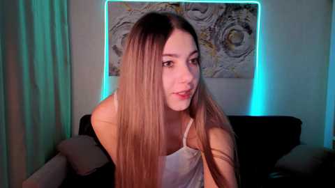 A video of a young Caucasian woman with long, straight, blonde hair, wearing a white spaghetti-strap top, sitting on a dark couch, with an abstract art piece and blue lighting in the background.