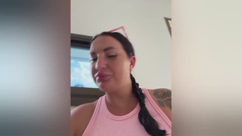 Video of a smiling, fair-skinned woman with dark hair braided, wearing a pink tank top, against a blurred, neutral-toned background.