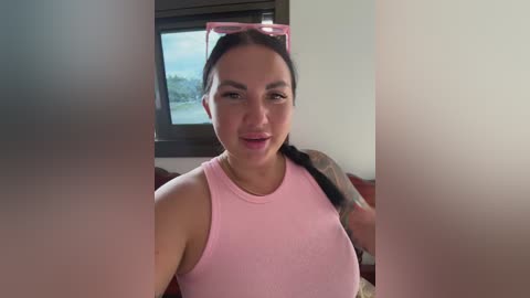 A video of a Latina woman with a medium complexion, dark hair, wearing a pink tank top, and a pink headband. She stands indoors with a blurred window behind her, revealing a green landscape.