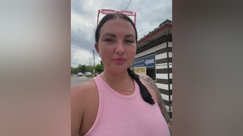 Video of a young woman with light skin, dark hair in a ponytail, wearing a pink tank top, standing in a cloudy outdoor setting, possibly a bus stop.