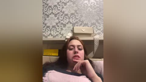 Video of a young woman with light skin and long brown hair, lying on a bed with a white patterned wall in the background. She has a contemplative expression, with one hand resting on her chin.