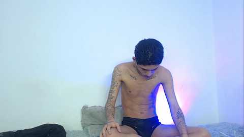 Video of a shirtless, tattooed, slender young man with short dark hair in black shorts, sitting on a gray couch, illuminated by blue and purple lights, against a plain white wall.