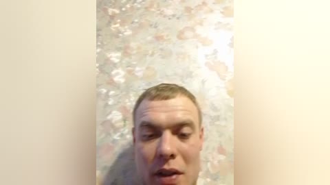 Video of a pale-skinned, balding man with a neutral expression, standing in front of a textured, floral-patterned wallpaper. The background is beige, and the image has a slightly blurry, amateur quality.