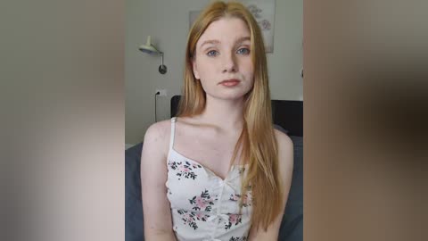 Video of a young, pale-skinned woman with long, straight red hair, wearing a floral-patterned white tank top, sitting on a bed in a minimalist room with neutral-colored walls and a white wall lamp.
