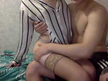 A video of a topless man and a woman in striped clothing, holding each other intimately on a bed with teal sheets and a grey wall.