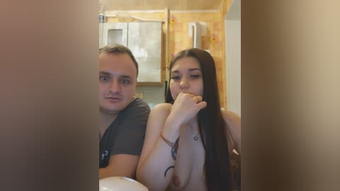 Video of a young couple, the man with short hair, wearing a grey t-shirt, and the woman with long brown hair, topless, in a bathroom with beige walls and a wooden door.