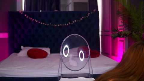 Video of a modern, dimly lit bedroom with a clear, round mirror placed on a nightstand, reflecting a plush, dark blue tufted headboard. Red pillows accent the white bed, while pink lighting creates a cozy ambiance.