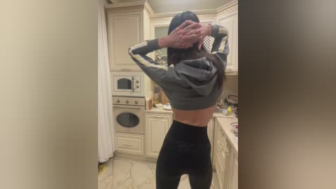 Video of a woman in a kitchen, seen from behind, wearing a grey crop top and black leggings. She has her hands in her hair, revealing a slender, toned physique. The kitchen has beige cabinetry and a tiled floor.
