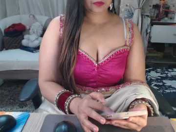 Video of a South Asian woman with medium skin tone and long, straight black hair, wearing a pink, sequined blouse and white saree, sitting at a desk with a computer and accessories, in a cozy, cluttered room.