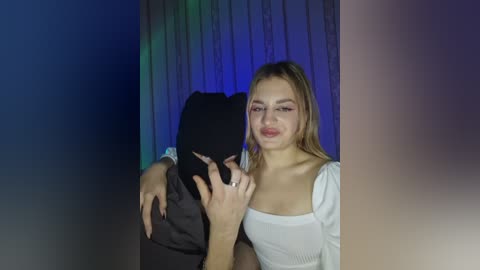 Video of a young Caucasian woman with long blonde hair, wearing a white off-shoulder top, hugging a masked person in a dark outfit. The background has a gradient of blue and purple hues.