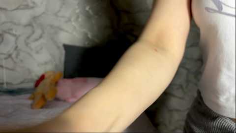 Video of a fair-skinned arm, wearing a white shirt, in a dimly lit room with grey marble-patterned wallpaper and a pink pillow.