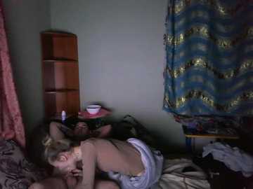 Video of a dimly lit bedroom scene featuring a man lying on his side, shirtless, with a woman leaning over him. The room has a wooden shelf, blue and gold-patterned curtain, and scattered clothes on the bed.