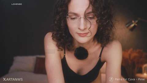 Video of a fair-skinned woman with curly black hair, wearing glasses and a black top, leaning forward in a dimly lit bedroom.