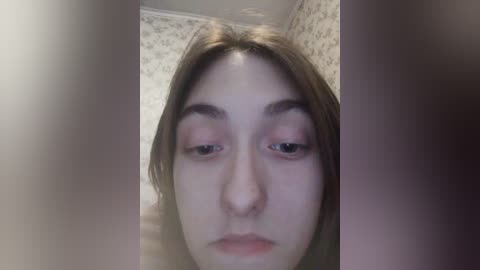 A close-up video of a young woman with fair skin, brown hair, and light pink eye makeup, set against a wallpapered background with a floral pattern. The image is slightly out of focus.