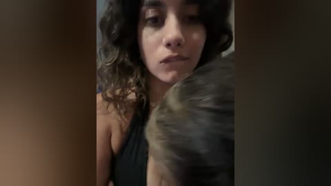 A video of a young woman with curly dark hair, wearing a black sleeveless top, hugging a man with a grey beard, in a dimly lit, blurry indoor setting.