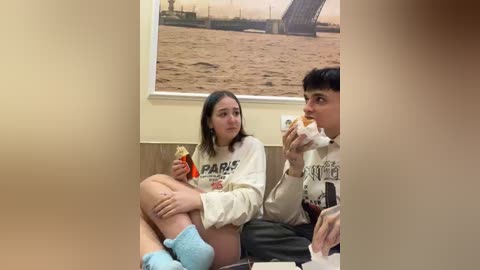 Video of two young people eating sandwiches in a modern office, with a large wall mural of a shipyard in the background.