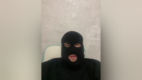 Video of a person wearing a black ski mask, sitting in a chair with a white cushion, against a textured gray wall. The person's facial features are obscured, and the scene appears dimly lit.