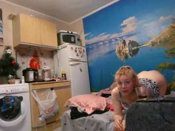 Video of a young woman with blonde hair, wearing glasses and a black top, bending over a bed in a small, cluttered room with a vibrant mural of a lake and mountains on the wall.