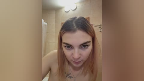 Video of a young Caucasian woman with shoulder-length, ombre hair in a beige-tiled bathroom. She has a tattoo on her left collarbone and is topless, with her facial expression showing slight surprise.