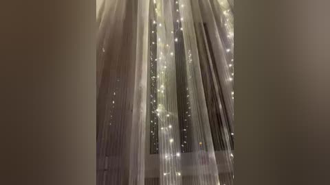Video of a window adorned with sheer white curtains and sparkling fairy lights, creating a whimsical, romantic ambiance. The background shows a blurred view of a dimly lit room, enhancing the soft, dreamy atmosphere.
