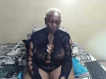 Video of an older black woman with short white hair, wearing a black lace bodysuit, sitting on a bed with a zebra-patterned sheet in a dimly lit room.