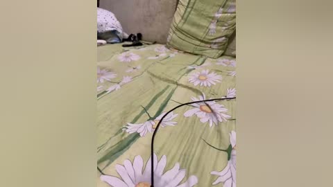 Video of a neatly made bed with a green floral sheet featuring white daisies and green stems. A beige pillow and a polka-dotted pillow are visible, and a pair of black shoes is placed on the floor beside the bed.