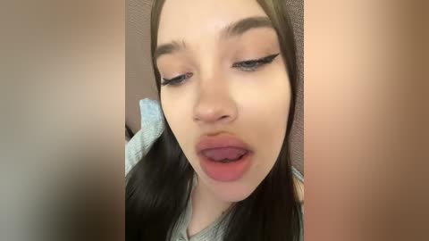 Video of a young woman with fair skin, dark hair, and long eyelashes. She has her mouth slightly open and her eyes half-closed, suggesting a seductive or playful expression. She wears a light-colored top. The background is blurred, featuring neutral colors.