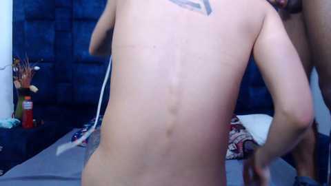 Video of a nude man with a slender physique and pale skin, standing in a dimly lit bedroom with a blue wall, a bed, and decorative items.