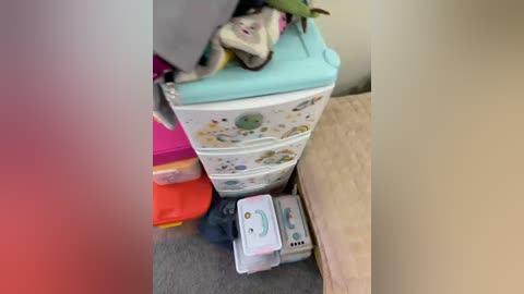 Video of a cluttered bedroom corner with a white toy chest, a beige bed, colorful storage bins, a small white electronic device, and a green plush toy atop the chest.