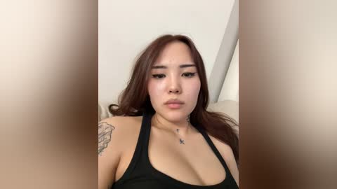 Video of an Asian woman with long, wavy brown hair, wearing a black halter top, showing a tattoo on her left shoulder. She has a neutral facial expression. The background is a blurred beige wall.