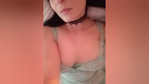 Video of a fair-skinned woman with dark hair, wearing a black choker and a green lace camisole, lying on a bed with a soft, peach-colored background.