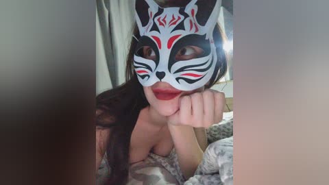 Video of a woman with fair skin, long dark hair, and red lipstick, wearing a white mask with black and red patterns, resting her chin on her hand, in a dimly lit bedroom with white curtains.