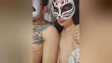 Video of a topless Asian woman with a fox mask and tattooed shoulders, standing close to a shirtless man with a beard, both indoors.