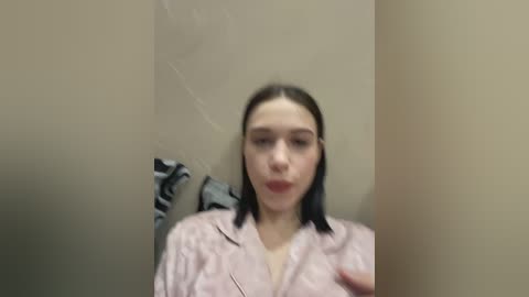 A video of a young woman with straight, dark hair, wearing a pink, patterned robe, standing in a beige-walled room. The image is slightly blurred, making details indistinct.