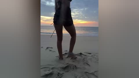 A video of a woman standing on a sandy beach at sunset, wearing a black, strapless swimsuit, with her back to the camera, capturing a serene, peaceful beach scene.